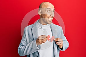 Bald man with beard holding broken heart paper shape smiling looking to the side and staring away thinking