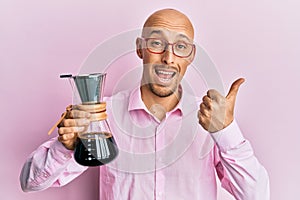 Bald man with beard holding coffee maker with filter pointing thumb up to the side smiling happy with open mouth