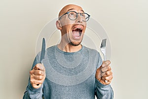 Bald man with beard holding fork and knife ready to eat angry and mad screaming frustrated and furious, shouting with anger