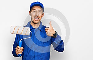 Bald man with beard holding roller painter smiling happy and positive, thumb up doing excellent and approval sign