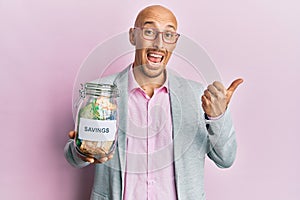 Bald man with beard holding savings jar with south african rands money pointing thumb up to the side smiling happy with open mouth