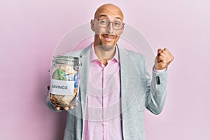 Bald man with beard holding savings jar with south african rands money screaming proud, celebrating victory and success very