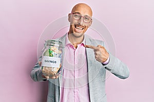 Bald man with beard holding savings jar with south african rands money smiling happy pointing with hand and finger