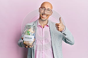 Bald man with beard holding savings jar with south african rands money smiling happy and positive, thumb up doing excellent and