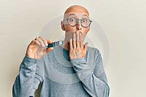 Bald man with beard holding ssd memory covering mouth with hand, shocked and afraid for mistake