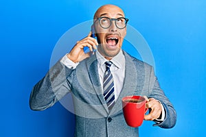 Bald man with beard using smartphone and drinking a cup of coffee angry and mad screaming frustrated and furious, shouting with