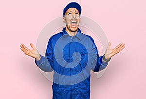 Bald man with beard wearing builder jumpsuit uniform crazy and mad shouting and yelling with aggressive expression and arms raised