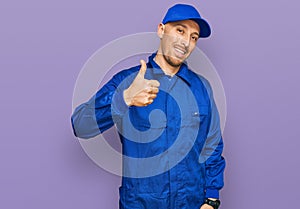 Bald man with beard wearing builder jumpsuit uniform doing happy thumbs up gesture with hand