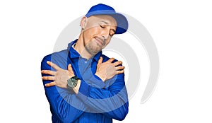 Bald man with beard wearing builder jumpsuit uniform hugging oneself happy and positive, smiling confident