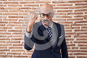 Bald man with beard wearing business clothes and glasses angry and mad raising fist frustrated and furious while shouting with