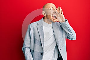 Bald man with beard wearing business jacket shouting and screaming loud to side with hand on mouth