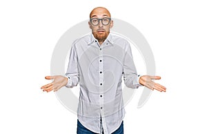 Bald man with beard wearing business shirt and glasses clueless and confused expression with arms and hands raised