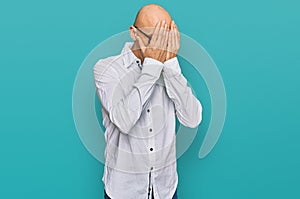 Bald man with beard wearing business shirt and glasses with sad expression covering face with hands while crying