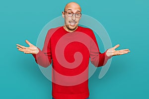 Bald man with beard wearing casual clothes and glasses clueless and confused expression with arms and hands raised