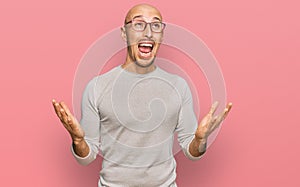 Bald man with beard wearing casual clothes and glasses crazy and mad shouting and yelling with aggressive expression and arms