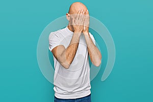 Bald man with beard wearing casual white t shirt with sad expression covering face with hands while crying