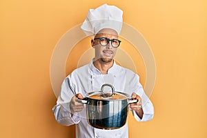 Bald man with beard wearing professional cook holding cooking pot clueless and confused expression