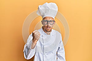 Bald man with beard wearing professional cook uniform angry and mad raising fist frustrated and furious while shouting with anger
