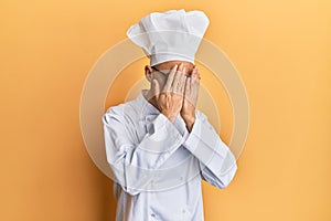 Bald man with beard wearing professional cook uniform with sad expression covering face with hands while crying