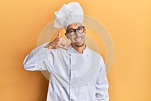 Bald man with beard wearing professional cook uniform smiling doing phone gesture with hand and fingers like talking on the