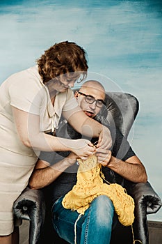 A bald man with a funny face and glasses obliquely knits while sitting on a wreck. Come on skillful man or handyman.