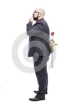 A bald man in glasses with a beard holds a red rose behind his back and looks up. Dressed in a formal suit. Full growth. Isolated