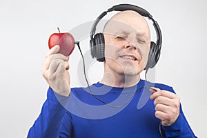 Bald man with headphones listens to music through a red apple player. Metaphor and concept of vitamin benefits in music and sound