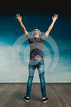 A bald man in headphones listens to music, standing with his back to the camera, raising his hands up on a blue background