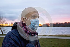 A bald man in a medical mask against the background of the setting sun and a pond. Concept: a violator of the quarantine