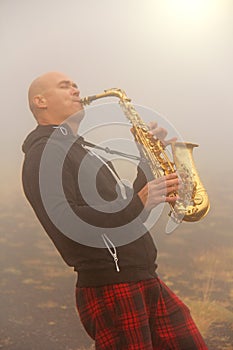 A bald man plays on a gold alto saxophone in the nature, against