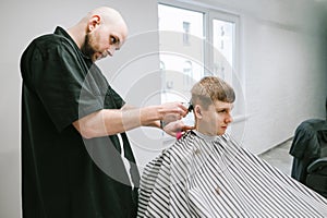 Bald man professional barber cuts the client`s hair with a clipper in the hands, looks at the young man`s head. A young man cuts