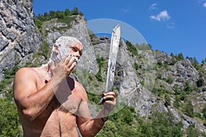 bald man shaves cheek with long knife phone on natural background. Morning on hike. Destination experience lifestyle concept