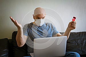 Bald man sitting in medical mask laptop with credit card in hand