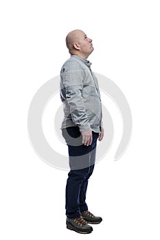 A bald man stands and looks up. A guy in a gray shirt and blue trousers. Full height. Side view. Isolated on a white background.