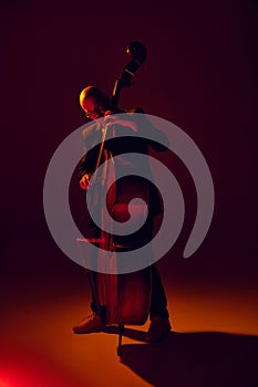 Bald man, talented bassist performing jazz compositions in red-yellow light against gradient studio background.