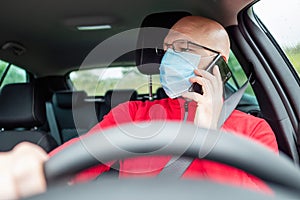 Bald man talking on his smart phone in a car, holding driving wheel and wearing red shirt and blue medical mask