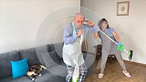 Bald man and woman in hair curlers in home clothes and aprons dance merrily with a vacuum cleaner, imitating playing the guitar,