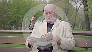 The bald mature man sitting in the park on the bench reading a book. Little boy coming from behind and putting hands on