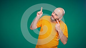 Bald middle-aged man with a beard in an orange t-shirt points with his hand to the up and left on a green background