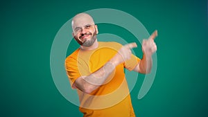 Bald middle-aged man with a beard in an orange t-shirt points with his hand to the up and right on a green background