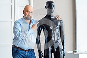 Bald serious adult man with beard, holding shoulders naked plastic black manikin