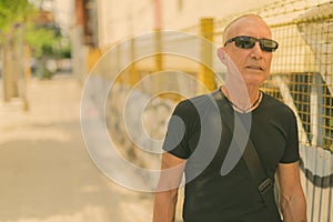 Bald senior tourist man thinking while wearing sunglasses against dirty urban wall with graffiti in the public street