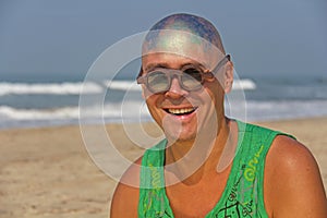 A bald and unusual young man, a freak, with a shiny bald head and round wooden glasses on the background of the beach and the sea