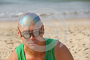 A bald and unusual young man, a freak, with a shiny bald head and round wooden glasses on the background of the beach and the sea