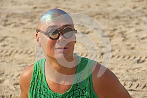 A bald and unusual young man, a freak, with a shiny bald head and round wooden glasses on the background of the beach and the sea