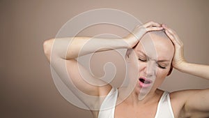 Bald woman holds her hands behind her head and screams. beige background, copy space