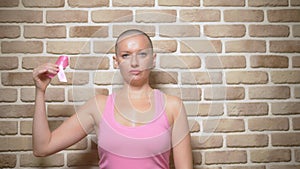 Bald woman Survivor after cancer proudly holds a pink ribbon in her hands against a brick wall.