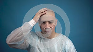 A bald young man with oncology sadly looks at the camera on a blue background. The patient touches his bald head. Hair