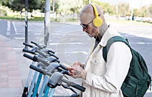 Bald young man in yellow headphones using a cell phone app to ride a public scooter in the city. Transportation, ecology and