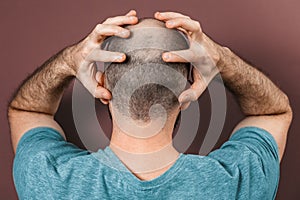 Baldy man grabs his head with his hands. Rear view. Brown background. The concept of alopecia and baldness
